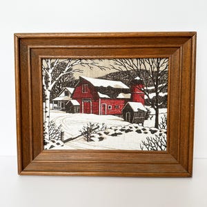 May include: Framed artwork depicting a winter scene with a red barn, snow-covered ground, and leafless trees. The artwork is set within a brown wooden frame, creating a rustic aesthetic. The scene evokes a sense of a rural winter landscape.