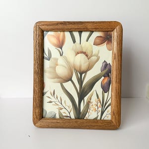May include: A framed floral print featuring watercolor-style tulips in cream and peach hues, with green leaves and accents of purple and yellow flowers. The print is set within a rectangular wooden frame with rounded corners.