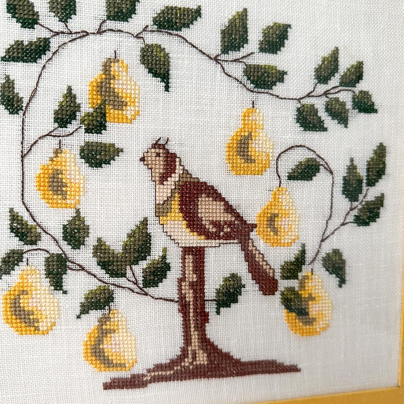 May include: Cross-stitch embroidery of a bird perched on a tree branch with yellow pears. The bird is brown, tan, and yellow, with a detailed design. The pears are yellow with white highlights. The leaves are dark green, and the background is white.