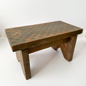 May include: A small, rectangular wooden stool with a checkerboard pattern on the top surface. The stool is stained a dark brown color, with the checkerboard squares in a darker shade of green. The legs are angled for stability.