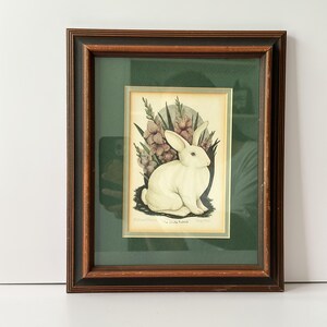 May include: Framed print featuring a white rabbit illustration with purple flowers. The artwork is set against a green background and framed in a dark brown wooden frame. The print is labeled "The White Rabbit".