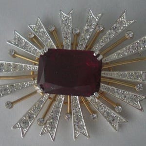 May include: A gold and silver brooch with a large, dark red, faceted gemstone in the center. The brooch is surrounded by smaller, clear rhinestones set in a starburst pattern.