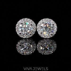 Pode incluir: Dois brincos de diamante com um halo de pedras menores, sobre um fundo preto. Os brincos são redondos e brilhantes, com corte brilhante. A marca "VIVA JEWELS" está na parte inferior.