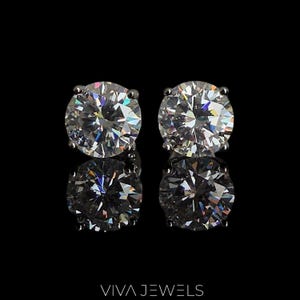 Puede incluir: Dos pendientes de botón redondos de diamantes transparentes con múltiples facetas, engastados en metal plateado. Los pendientes se muestran sobre una superficie negra reflectante, con la marca "VIVA JEWELS" en la parte inferior.