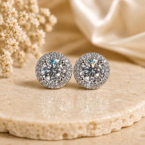 May include: A pair of round, silver-toned stud earrings with a large, clear center stone surrounded by smaller, sparkling stones. The earrings are set on a light-colored, textured surface, with a soft, blurred background.