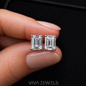 May include: A pair of emerald-cut diamond stud earrings. The earrings are set in a silver metal and are held against a black background. The earrings are from the Viva Jewels brand.