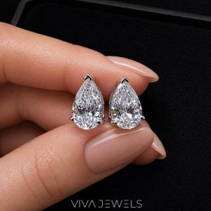 May include: A pair of pear-shaped diamond stud earrings. The earrings are set in silver metal and are held by a hand with a neutral skin tone. The earrings are sparkling and reflect light. The brand name "VIVA JEWELS" is at the bottom of the image.