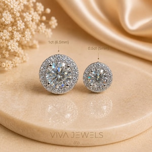 May include: Two diamond stud earrings, one 6.5mm and one 5mm, with a halo of smaller stones. The earrings are set on a neutral-toned surface with the text "VIVA JEWELS" at the bottom.