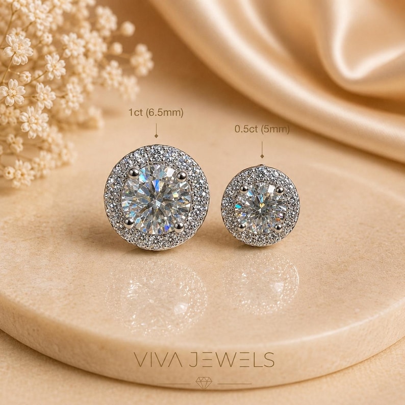 May include: Two diamond stud earrings, one 6.5mm and one 5mm, with a halo of smaller stones. The earrings are set on a neutral-toned surface with the text "VIVA JEWELS" at the bottom.