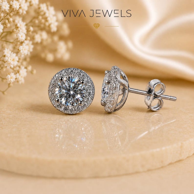 May include: A pair of silver stud earrings with a halo of small, sparkling stones surrounding a larger, clear center stone. The earrings are displayed on a neutral surface with the text "VIVA JEWELS" at the top.