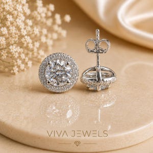 May include: A pair of sparkling diamond stud earrings with a halo of smaller diamonds. The earrings are set in a silver metal and are displayed on a neutral-toned surface. The brand name "VIVA JEWELS" is visible at the bottom.