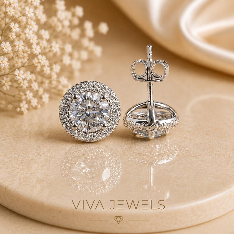 May include: A pair of sparkling diamond stud earrings with a halo of smaller diamonds. The earrings are set in a silver metal and are displayed on a neutral-toned surface. The brand name "VIVA JEWELS" is visible at the bottom.
