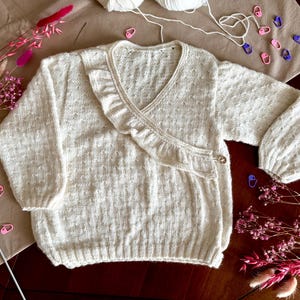 Peut inclure: Un pull cache-cœur tricoté de couleur crème avec une encolure en V ornée d'un volant sur le bord. Le vêtement possède des manches longues avec des poignets côtelés et une fermeture à bouton unique sur le côté. Le tricot présente un motif texturé.