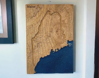 May include: Wooden wall art of the state of Maine, featuring a detailed relief map. The land is a light brown color, and the coastal areas are filled with a deep blue resin. The word "Maine" is carved at the top.