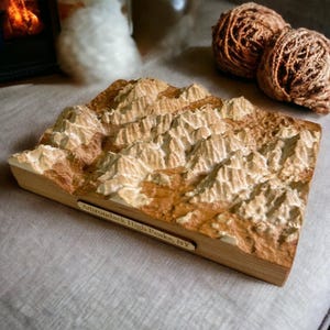 May include: A wooden relief map of the Adirondack High Peaks, NY. The map is carved with detailed mountain ridges and valleys, painted in shades of brown and cream. The base is a light wood, and a small label reads "Adirondack High Peaks, NY."
