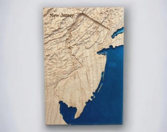 May include: A wooden relief map of New Jersey, featuring a detailed topographic representation. The land is a light tan color, with the ocean represented in a deep blue resin. The text "New Jersey" is carved into the wood.