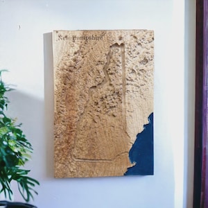 May include: A wooden wall art piece depicting the state of New Hampshire. The state's outline is carved into the wood, with the text "New Hampshire" at the top. The bottom right corner is painted blue.