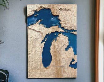 May include: Wooden wall art featuring a detailed map of Michigan with blue resin representing the Great Lakes. The word "Michigan" is engraved at the top. The artwork is mounted on a light-colored wooden surface.