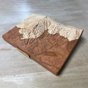 May include: A wooden relief carving of a mountain range, featuring a light-colored peak and a darker, reddish-brown base. The carving is mounted on a rectangular wooden base, with the text "Killington, VT" visible on a small wooden tag.