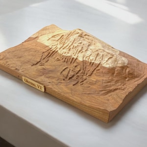 May include: A wooden Okemo, VT topographical map. The map is carved from light brown wood, with a lighter area representing the mountain's peak. A small rectangular plaque with the text "Okemo, VT" is attached to the front.