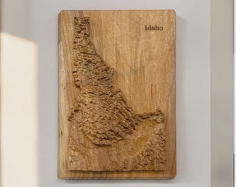 May include: A wooden wall art piece featuring a detailed relief map of Idaho. The state's outline is carved into the light brown wood, with the word "Idaho" engraved at the top. The piece is rectangular and mounted on a wall.