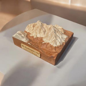 May include: A wooden relief sculpture of Mt. Washington, NH, part of the Northern Presidential range. The sculpture features detailed peaks in a light beige color, set against a brown and tan landscape, mounted on a wooden base. The name is inscribed on a small plaque.