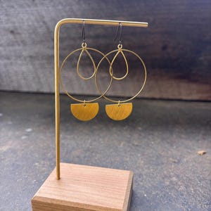 May include: Gold-toned earrings with a circular design, teardrop accents, and half-moon shapes. The earrings are displayed on a gold stand with a wooden base, showcasing their modern aesthetic. The earrings are a stylish accessory.