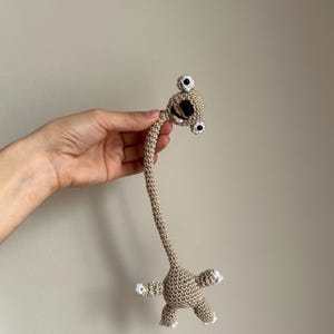 May include: A handmade crochet creature in tan with a long neck, small body, and four limbs. The creature has two large eyes and a small mouth. The tips of the limbs are white.
