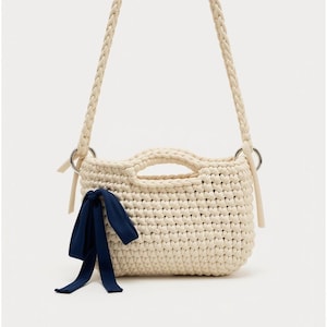 Può includere: Una borsa all'uncinetto color crema con una spessa tracolla intrecciata fissata da anelli in metallo argentato. La borsa ha un corpo rettangolare strutturato con un'apertura per il manico superiore e un nastro in tessuto blu scuro sul davanti.
