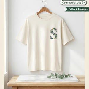Floral embroidery font mockup on cream t-shirt hanging on wooden hanger against white wall. Botanical letter S on left chest with teal green vines, yellow and pink blooms. Oak table with marble slab and eucalyptus below. Commercial use OK, Full A-Z included. 26 letters, 6 sizes 3.5 to 6 inch, 10 formats. PES DST JEF XXX VP3 VIP HUS EXP ART SEW.