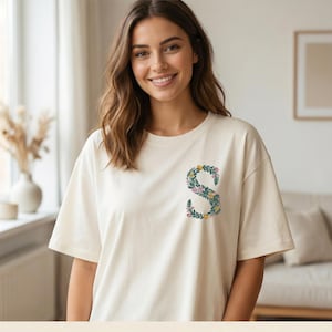 Floral embroidery font lifestyle mockup. Smiling young woman wearing cream oversized t-shirt with botanical letter S on left chest — teal green vines, yellow and pink blooms. Bright airy home interior background. Commercial use OK. 26 letters, 6 sizes 3.5 to 6 inch, 10 formats. PES DST JEF XXX VP3 VIP HUS EXP ART SEW.