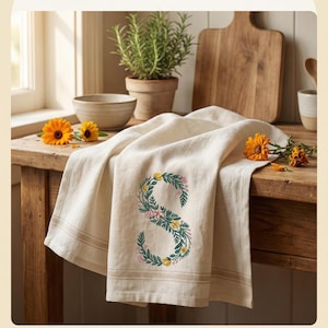 Floral embroidery font mockup on cream linen kitchen towel draped over rustic wooden table. Botanical letter S with teal green vines, yellow and pink blooms. Warm farmhouse kitchen scene with potted rosemary, wooden cutting board and marigold flowers. Commercial use OK. 26 letters, 6 sizes 3.5 to 6 inch, 10 formats. PES DST JEF XXX VP3 VIP HUS EXP ART SEW.