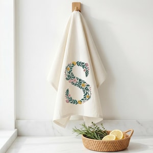 Floral embroidery font mockup on cream kitchen towel hanging on wooden wall hook against white wall. Botanical letter S with teal green vines, yellow and pink blooms. Wicker basket with fresh lemons and herbs on marble surface below. Commercial use OK. 26 letters, 6 sizes 3.5 to 6 inch, 10 formats. PES DST JEF XXX VP3 VIP HUS EXP ART SEW.