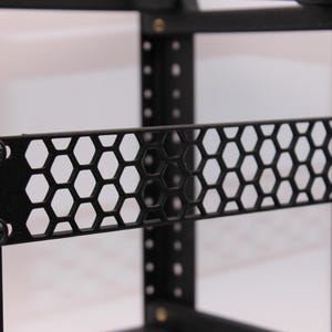 May include: Close-up of a black metal rack component with a honeycomb pattern. The component is secured with black screws and is part of a larger rack structure. The design features a series of hexagonal cutouts.