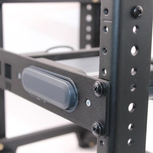 May include: Close-up of a black metal rack system with multiple shelves and support beams. A gray, rectangular button is mounted on the front of the rack. The rack is secured with black screws and has several holes for adjustments.