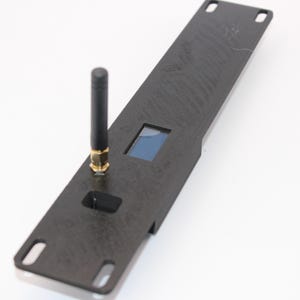 May include: A black rectangular electronic device with a small blue screen and an antenna. The device has a textured surface and several cut-out sections. The antenna is black with a gold-colored base.