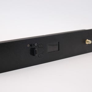 May include: A black rectangular electronic component with a USB port, small screen, and a gold-colored connector. The component has mounting holes on each end and is set against a white background.