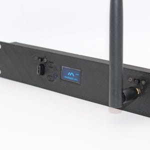 May include: A black electronic device with a screen displaying "IN -" and "medusa.net". It has a long antenna and several ports. The device is rectangular with mounting holes on each end.