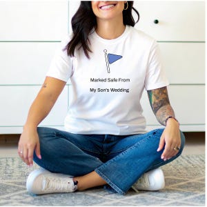 Puede incluir: Camiseta blanca con un diseño de bandera azul y el texto "Marked Safe From My Son's Wedding". La camiseta está hecha de un material suave y es adecuada para ropa casual.