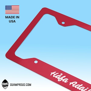 May include: Red license plate frame with the text "Håfa Adai" in white script. The frame has a rectangular shape with rounded corners and mounting holes. The American flag and "MADE IN USA" are in the upper left corner. The website GUAMPROUD.COM is also visible.