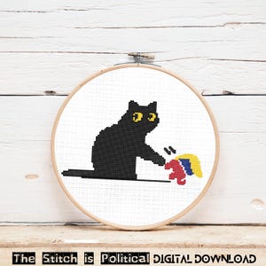 Peut inclure: Modèle de point de croix d'un chat noir aux yeux jaunes qui donne un coup de patte à un objet rouge, jaune et bleu. Le motif est dans un cercle de broderie en bois.