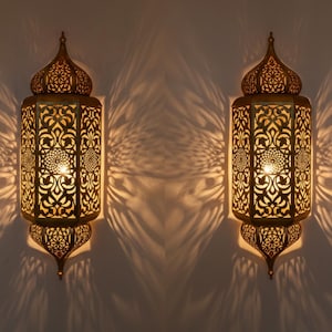 Peut inclure: Deux appliques murales dorées ornées de motifs ajourés complexes. Les appliques ont une forme de lanterne traditionnelle, avec un sommet et un bas pointus. La lumière intérieure projette des ombres décoratives sur le mur, créant une ambiance chaleureuse.