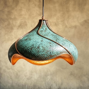 May include: A unique, handcrafted pendant light fixture. The lamp features a textured, teal-colored shade with copper accents and a warm, golden interior. The fixture hangs from a black cord, showcasing an elegant, artistic design.