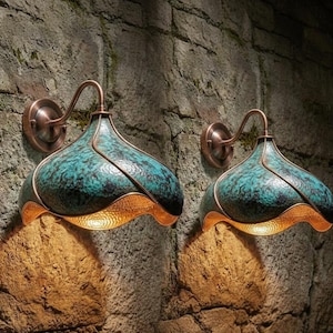 Op de afbeelding: Twee teal en koperen wandkandelaars zijn bevestigd aan een stenen muur. De wandkandelaars hebben een uniek, gebogen ontwerp met een getextureerde teal bovenkant en een koperkleurige onderkant. De wandkandelaars zijn verlicht en werpen een warme gloed op de steen.
