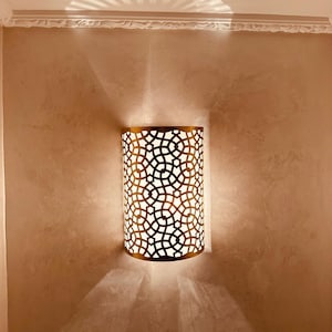 Può includere: Un'applique da parete dorata con un motivo geometrico. L'applique è accesa e proietta un'ombra sul muro.