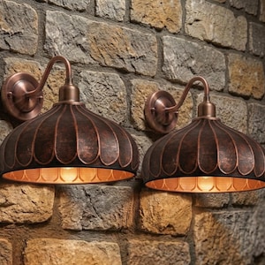 Pode incluir: Duas arandelas de parede em estilo antigo com acabamento em cobre. Cada arandela apresenta uma cúpula com ranhuras verticais e um braço curvo. As arandelas são montadas em uma parede de pedra texturizada.