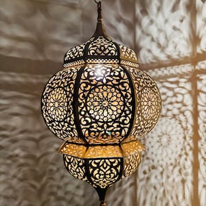 May include: An ornate, hanging metal lantern with intricate cut-out patterns. The lantern has a bulbous centre and a pointed top and bottom. The warm light inside casts decorative shadows on the wall. The lantern is suspended from a chain.