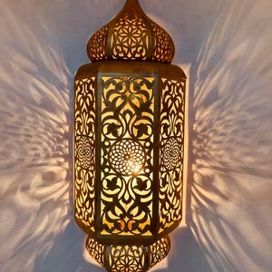 May include: A gold metal wall sconce with intricate cut-out designs. The sconce has a dome-shaped top and a rectangular body. The light shines through the cut-outs, creating a beautiful pattern on the wall.
