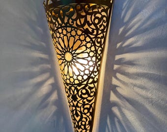May include: A decorative wall sconce with a detailed, openwork design. The sconce is shaped like a cone, with intricate patterns and a warm light emanating from within. The sconce is made of a dark metal and is mounted on a light blue wall.
