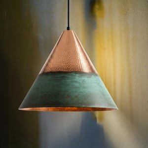 Puede incluir: Una lámpara colgante de cobre con forma cónica. La parte superior tiene una textura martillada y un color cobre cálido, mientras que la sección inferior es de color verde desgastado. Cuelga de un cable negro.